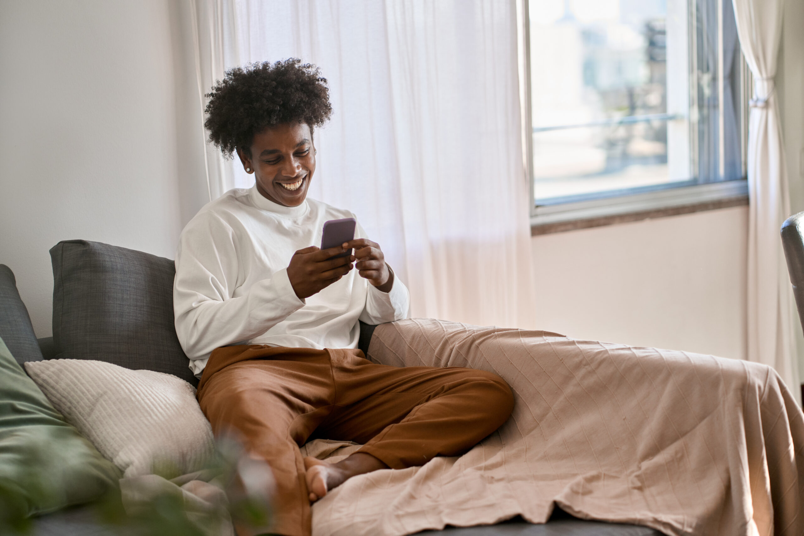 Happy relaxed gen z African American teen sitting on couch at home holding smartphone, using cell phone mobile apps, shopping or playing game. Social media influence concept. Authentic shot.